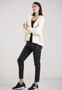 Cremefarbener gestrickter Cardigan über einem schwarzen Oberteil, kombiniert mit schwarzen Kunstlederhosen und schwarzen Sneakers mit silbernen Akzenten, die eine lockere Passform zeigen.