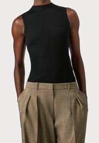 En person bär en tajt svart ärmlös topp och högmidjade beige veckade byxor, med händerna i fickorna, mot en enkel bakgrund.