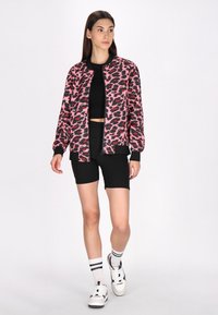 Giacca bomber con stampa leopardata rosa e nera, chiusura con zip, polsini a costine, abbinata a un top corto nero e pantaloni corti, completata con sneakers.