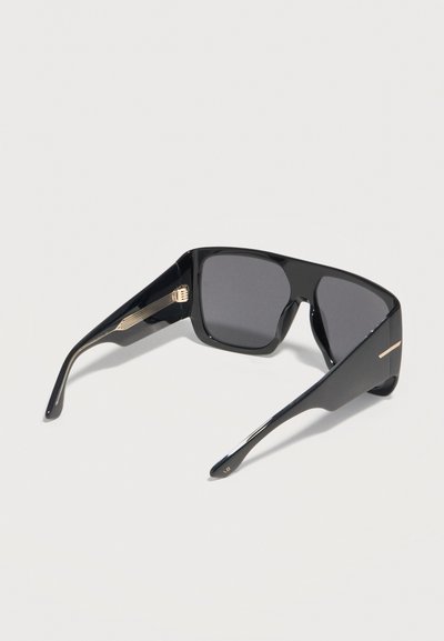 Μαύρα υπερμεγέθη τετράγωνα γυαλιά ηλίου με σκούρους φακούς και χρυσές λεπτομέρειες στα μπράτσα, εμφανιζόμενα πάνω σε απλό φωτεινό φόντο.