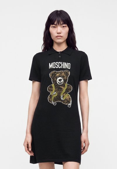 Жена с черна поло рокля Moschino с графика на плюшено мече, увито в жълта метър лента отпред.