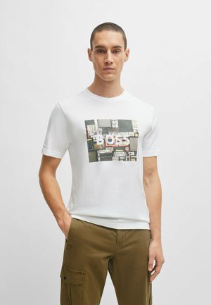 Camiseta blanca de manga corta con un estampado gráfico que presenta el texto "BOSS" en el centro, combinada con pantalones cargo color beige.