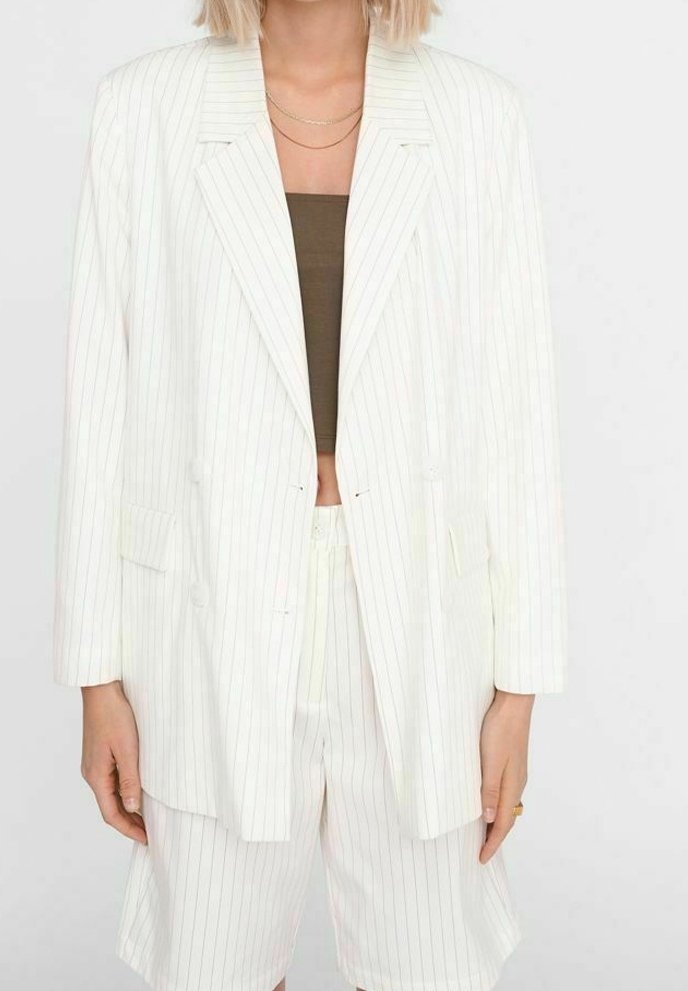 Femme portant un blazer blanc à fines rayures et un short assorti avec un crop top marron et des colliers dorés superposés.