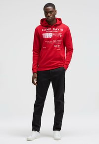 Red hoodie with white graphic text, drawstring closure, and kangaroo pocket, paired with black trousers and white sneakers.