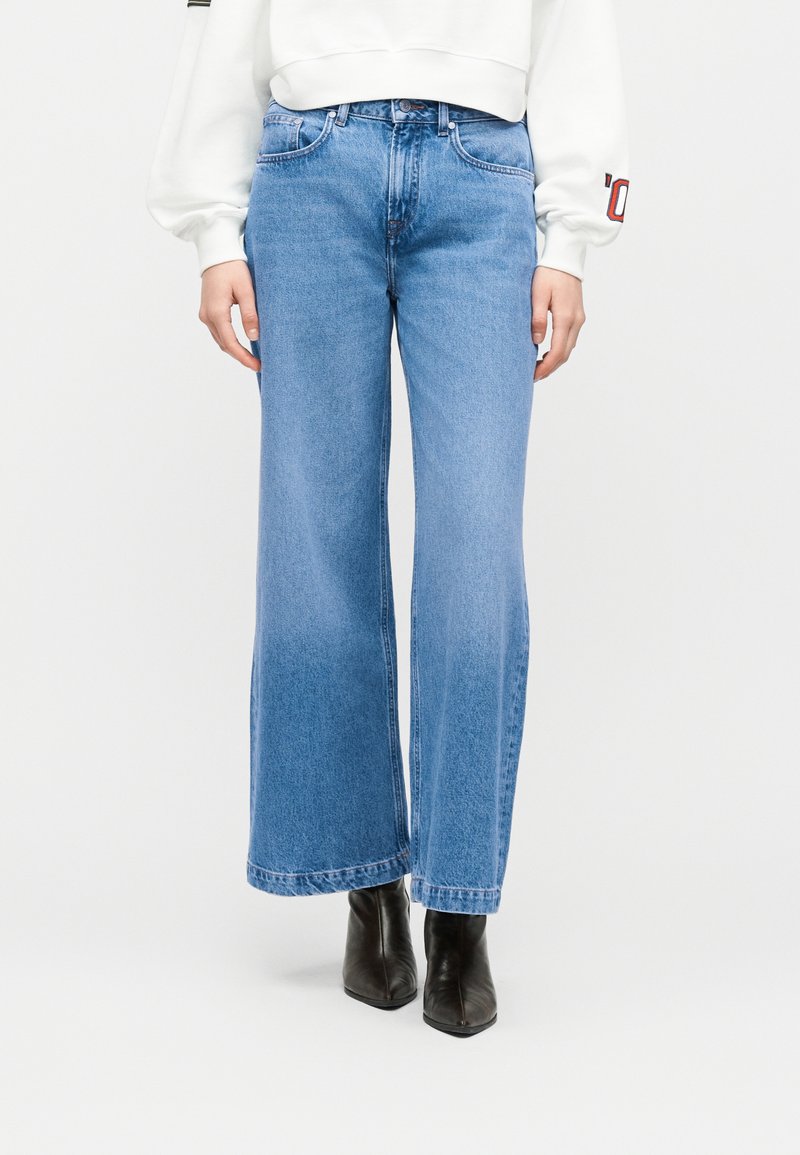 Άτομο που φοράει ανοιχτό μπλε τζιν με φαρδύ πόδι, λευκό φούτερ και μαύρα μποτάκια με μυτερό δάχτυλο, στέκεται μπροστά σε απλό φόντο.