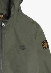 Olive zielona kurtka z kapturem z zamkiem z przodu, mały okrągły logo na klatce piersiowej i kwadratowa naszywka z logo na lewym rękawie.