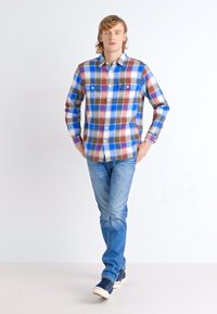 Geruite shirt met blauwe, rode, bruine en witte vierkanten, met knoopsluiting kraag, borstzakken, gedragen met lichtblauwe jeans en sneakers.