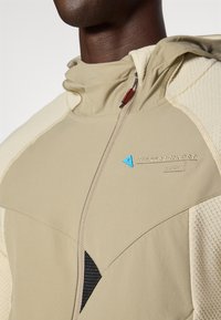Casaco desportivo bege com acentos texturizados em creme, fecho de zip e um logotipo em triângulo azul. Apresenta um capuz e detalhe em malha preta.