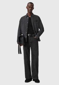 Uomo alto che indossa una camicia nera a bottoni sopra una maglietta nera, jeans neri, cintura nera, scarpe nere, mentre tiene una borsa di pelle nera su uno sfondo grigio chiaro.