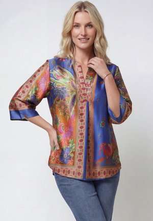 Blond meisje met blauwe spijkerbroek en een kleurrijk bloemetjesblouse, glimlachend met één hand op de heup en de andere bij haar nek.