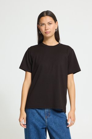 Jeune femme aux longs cheveux foncés portant un T-shirt noir uni et un jean bleu, debout devant un fond gris clair.