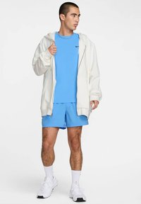 Haut zippé léger blanc, T-shirt bleu et shorts bleus. Le modèle porte des chaussures de sport blanches et des shorts, mettant en avant une coupe décontractée et un contraste de couleurs.