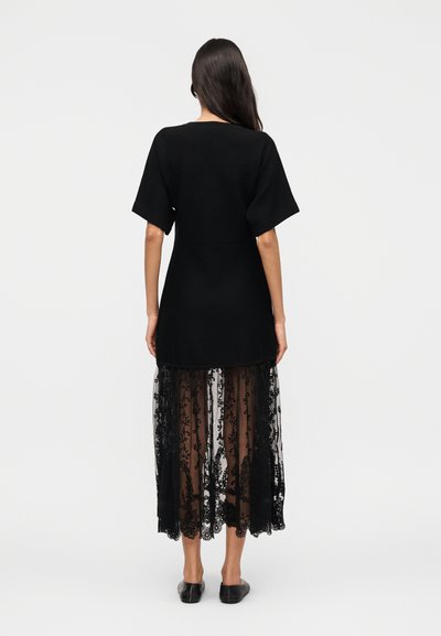 Γυναίκα με μακριά σκούρα μαλλιά που φοράει μαύρο φόρεμα με κοντά μανίκια και διαφανή φλοράλ δαντέλα στην φούστα, στέκεται γυρισμένη με την πλάτη σε λευκό φόντο.