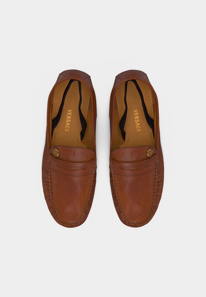 Coppia di mocassini Versace in pelle marrone con punte arrotondate, cuciture mocassino e dettaglio con bottone Medusa dorato su sfondo bianco.