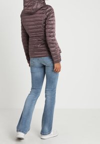 Padded, kaptur z miękkiego brązu z pikowanym wzorem, noszony na jasnoniebieskich dżinsach dzwonach i białych sneakersach, widok z tyłu.