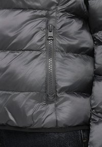 Τμήμα μαύρου μπουφάν puffer με σχέδιο καπιτονέ, που διαθέτει ανθεκτικό φερμουάρ και λείο, γυαλιστερό ύφασμα.