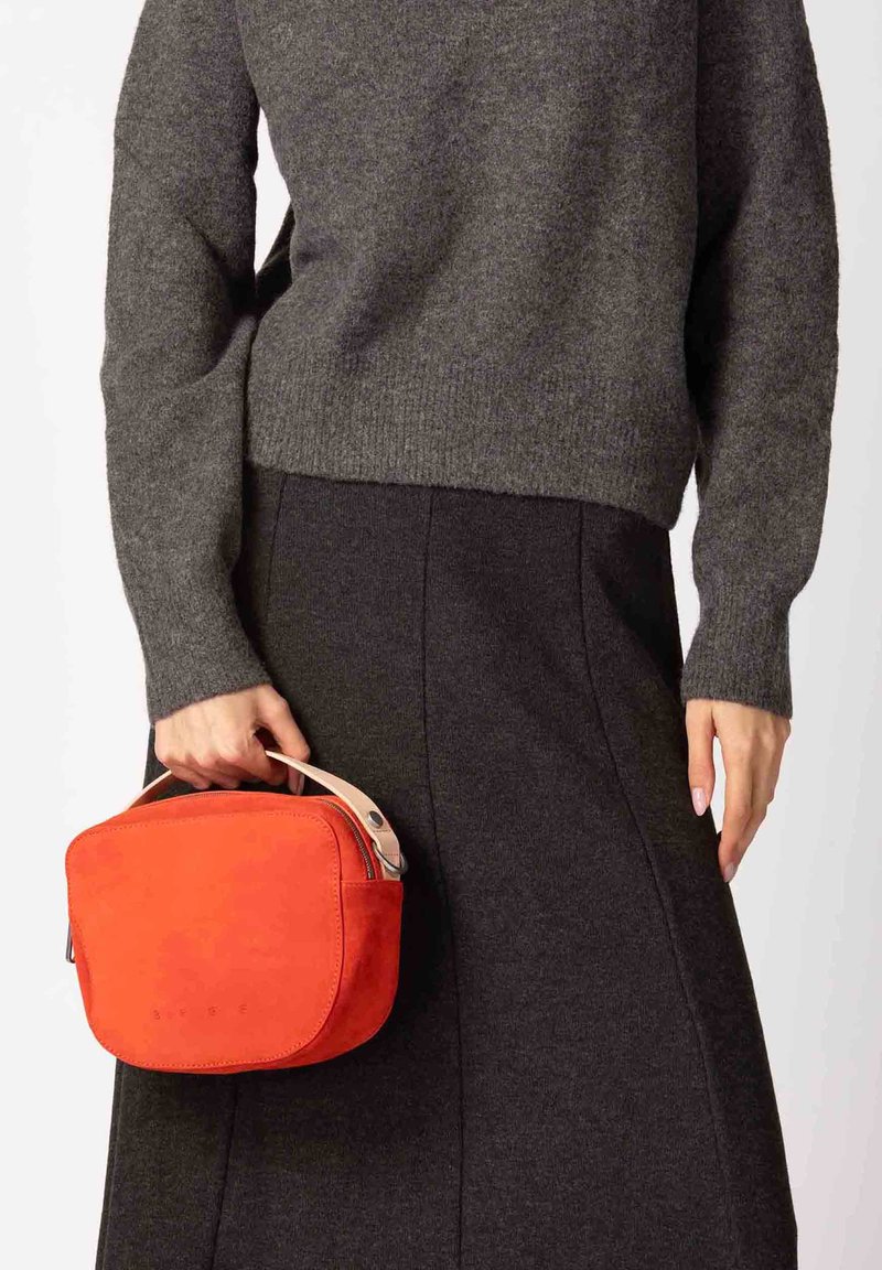 Rode suède handtas met een afgeronde vorm, leren handvat en ritssluiting. Draagt een grijze trui en een donkere rok.