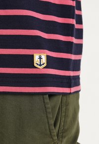 Navy and pink striped cotton t-shirt featuring a small anchor logo patch on a shield at the hem. Pairing with olive green pants.