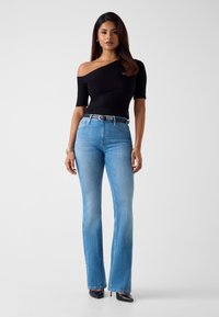 Schwarzes Off-Shoulder-Oberteil kombiniert mit hellblauen, ausgestellten Jeans. Die Jeans haben eine normale Taille und einen dunklen Gürtel, ergänzt durch spitze schwarze Schuhe.