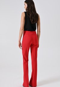Donna con lunghi capelli castani, che indossa un top senza maniche nero e pantaloni rossi a vita alta a campana, in piedi su uno sfondo chiaro e semplice.
