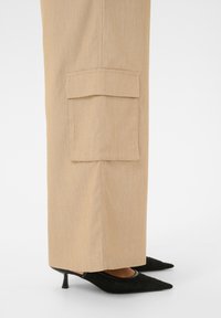 Pantalons larges beiges avec une poche latérale, associés à des talons aiguilles noirs à bout pointu, avec un dessus texturé et un petit talon stiletto.