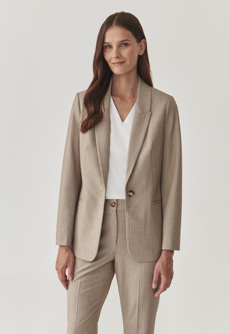 Completo beige con righe sottili, giacca su misura con chiusura a un bottone e pantaloni abbinati; tessuto texture, finitura morbida, sotto una maglietta bianca a scollo a V.