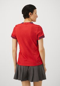Red athletic shirt with vertical ribbed texture, short sleeves featuring blue patterned accents. Paired with a gray pleated skirt.
