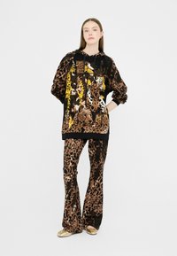 Ensemble de sweat à capuche et pantalon en imprimé léopard brun et noir, avec des graphismes animaliers détaillés et une coupe décontractée. Accents dorés sur les chaussures.