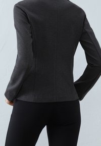 Chaqueta ajustada de color gris oscuro con mangas largas, textura suave, detalles de costuras a lo largo de la espalda y silueta entallada. Se lleva sobre pantalones negros.