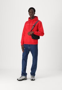 Raudona gobtuvė su "TOMMY JEANS" logotipu, derinama su mėlynomis džinsinėmis kelnėmis ir pilkais sportiniais batais. Juoda per petį nešama rankinė, stovinti ant paprasto fono.