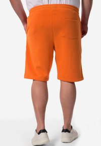 Oranžové bavlněné šortky s elastickým pasem, boční kapsou a zadní kapsou. Hladká textura a design až ke kolenům. Nosené s bílými teniskami.