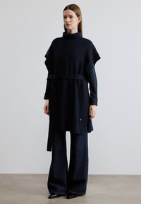 Πλεκτό ποντσο σε ναυτικό μπλε με ψηλό γιακά και φαρδιά μανίκια, σφιγμένο στη μέση με ζώνη που ταιριάζει. Συνδυασμένο με σκούρο παντελόνι φαρδιάς γραμμής.