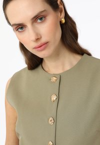 Haut sans manches à col en tissu vert olive, doté de boutons rehaussés d'or sur le devant et d'une finition lisse et texturée.