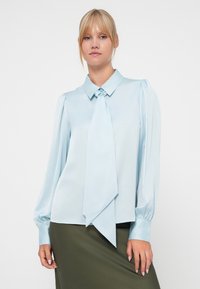 Blusa azul clara de cetim, com um laço no colarinho, mangas longas com puf e punhos com botão. Textura suave e design elegante.