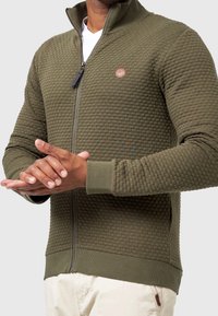 Homme portant une veste zippée texturée vert olive avec poignets et ourlet côtelés, et un pantalon blanc sur un fond uni.