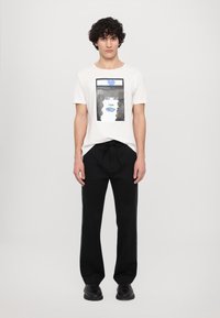 White t-shirt featuring a graphic print of a face and the text "PETULIA." Paired with black pants with green striping and a drawstring waist.