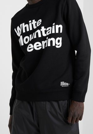 Sweat-shirt noir avec un grand texte blanc "White Mountaineering" et un petit texte imprimé près de la taille, porté avec un pantalon foncé.