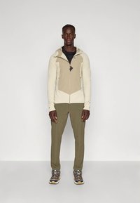 Vīriešu sporta apģērbs: bēša ar rāvējslēdzēju kapuce ar ģeometrisku dizainu, kopā ar olīvu zaļām biksēm un brūnām pārgājienu kurpēm ar baltiem akcentiem.