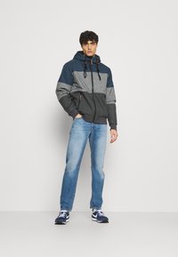 Color-blocked hooded jacket in dark blue, gray, and charcoal with front pockets, adjustable drawstrings, and a zip-up closure, paired with light blue jeans.