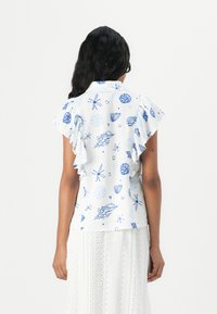 Hvit bluse med blå sjø-tema trykk, med volanger på ermene og krage. Stoffet ser lett ut og har en jevn tekstur.