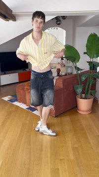Rumena in bela črtasta kratka majica s kratkimi rokavi, temni denim kratki hlače z raztrganimi robovi in srebrni športni čevlji, stoji v notranjosti z rastlinami.