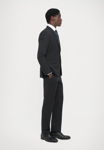 Mørk skræddersyet jakkesæt med slim fit, der har en jagtlapel, parret med en hvid skjorte og et mønstrede blå slips, suppleret med sorte sko.