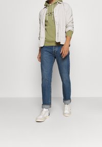 Helles beiges Jackett, olivgrüner Hoodie, blaue Jeans und weiße Sneaker. Die Jeans sind an den Säumen aufgerollt und haben eine lässige Passform.