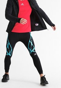 Persona con leggings de compresión negros y verde azulado, una camiseta roja de Adidas, zapatos negros y una chaqueta negra abriendo la chaqueta.