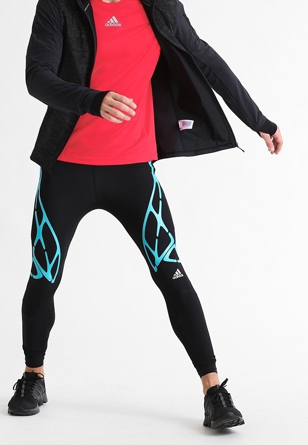 Persona con leggings de compresión negros y verde azulado, una camiseta roja de Adidas, zapatos negros y una chaqueta negra abriendo la chaqueta.