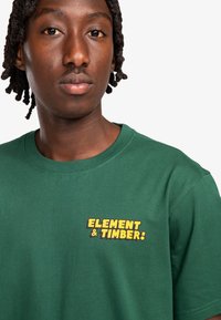Green cotton t-shirt featuring a yellow graphic text "ELEMENT & TIMBER!" on the left chest area. Short sleeves and a crew neckline.