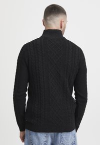 Schwarzer gestrickter Pullover mit hohem Rippkragen, der ein strukturiertes Kabelstrickmuster auf dem Rücken sowie Rippbündchen und -saum hat.
