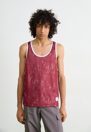 Top sin mangas de patrón rojo con un borde blanco, con una tela texturizada y una pequeña etiqueta con el logo cerca del dobladillo. Combinado con pantalones cargo grises.