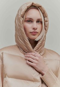 Beige puffer vest with a hood, featuring quilted sections and a smooth texture. Model's hand rests on the collar, wearing a light knit sweater.