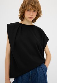 Top senza maniche nero con un design drappeggiato, caratterizzato da una spalla asimmetrica e una texture liscia. Abbinato a pantaloni in denim larghi.
